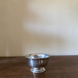 May include: A small, polished silver-toned metal bowl with a smooth, rounded body and a flared rim. The bowl features a circular pedestal base that elevates the main vessel, creating a classic, simple silhouette with a reflective metallic finish.