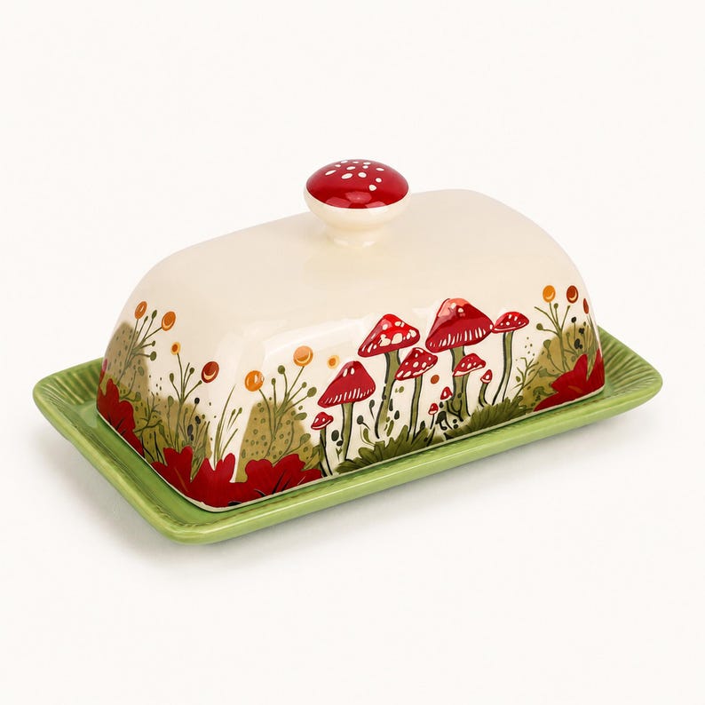 May include: A ceramic butter dish with a cream-colored lid featuring a red mushroom and green foliage pattern, topped with a red mushroom-shaped knob. The base is a rectangular, ribbed green plate with a raised edge for holding the lid securely.