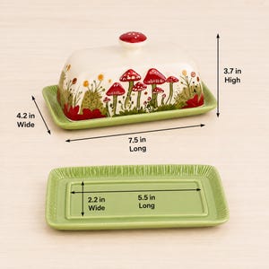 May include: A ceramic butter dish with a lid featuring red mushroom illustrations on a cream background. The lid is 7.5 inches long, 4.2 inches wide, and 3.7 inches high. The green base tray measures 5.5 inches long and 2.2 inches wide.