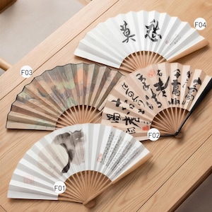 Pode incluir: Quatro leques de papel decorativos com cabos de madeira são exibidos em uma superfície de madeira. Os leques apresentam vários designs, incluindo caligrafia e pinturas em aquarela. Um leque tem uma ilustração de gato preto.