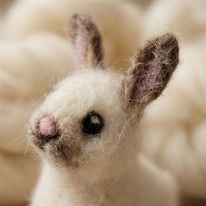 May include: A close-up of a handmade white bunny figurine with a pink nose and inner ears. The bunny has dark eyes and brown-tipped ears. The soft, fluffy texture suggests it is made of wool or a similar material.
