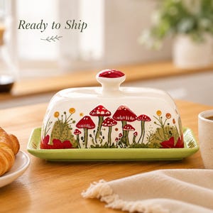 May include: A ceramic butter dish with a rectangular light green base and a white lid featuring a red mushroom pattern and a matching mushroom-shaped knob. The text Ready to Ship is printed in the upper left corner above a small leaf graphic.