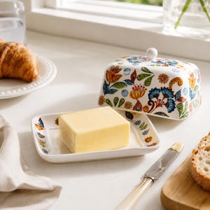 May include: A rectangular ceramic butter dish with a white base and a matching lid featuring a colorful floral and paisley pattern in shades of blue, orange, green, and red. The lid has a small white knob handle on top, and a block of yellow butter sits inside.
