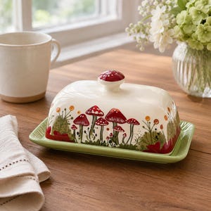 May include: A rectangular ceramic butter dish with a cream-colored lid featuring a hand-painted pattern of red mushrooms and green foliage. The lid has a round red knob on top, and the base is a solid light green with a raised rim edge.