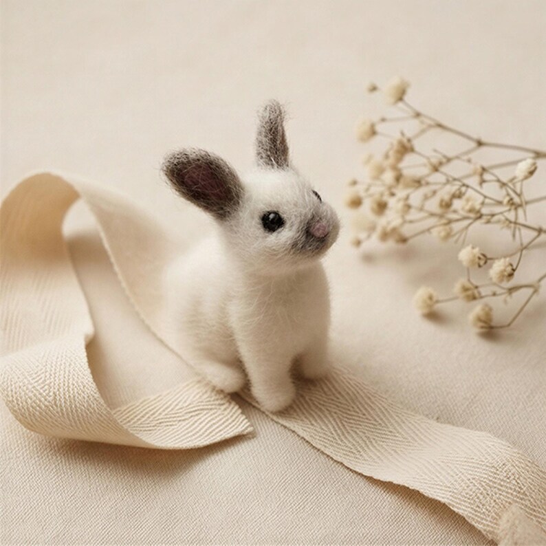 May include: A handmade white felt rabbit with grey-tipped ears and a grey nose sits on a cream-coloured ribbon. The rabbit has black eyes and is positioned next to delicate, dried flowers. The overall aesthetic is soft and natural.