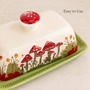 May include: A cream-colored ceramic butter dish with a red mushroom-shaped knob and a green ridged base. The lid features a painted forest scene with red mushrooms, green foliage, and orange berries. The text "Easy to Use" is printed on the lid.