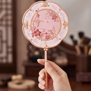 May include: A pink and gold hand fan with a floral and butterfly design. The fan has a round, ornate head with a detailed pattern and a long, slender handle. The fan is held by a person's hand.