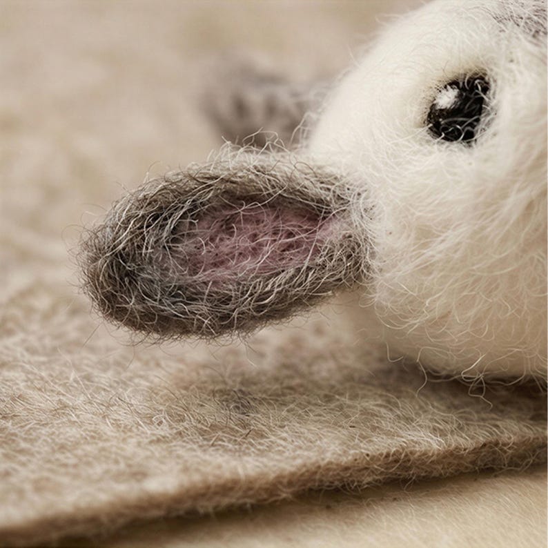 May include: Close-up of a handmade felted bunny. The bunny has a white face with a black eye and a grey ear with a pink interior. The bunny is lying on a beige felt surface.