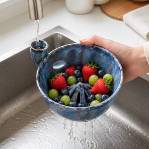 May include: A blue ceramic berry colander bowl with a reactive glaze finish, featuring a central raised cone for rinsing fruit and a small side handle. The bowl is filled with fresh red strawberries and green grapes while water flows through it.