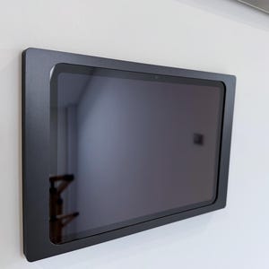 Puede incluir: Una tableta negra montada en una pared blanca. La tableta es rectangular con esquinas redondeadas y un marco negro. La pantalla es oscura, reflejando el entorno.