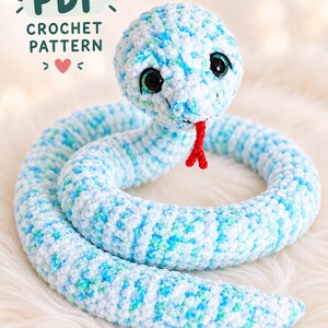May include: A crochet snake toy made of soft, variegated yarn in shades of white, light blue, and turquoise. The snake has large, dark, glossy eyes and a small, red, bifurcated tongue. Text in the upper left corner reads PDF CROCHET PATTERN.