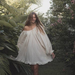 May include: An off-the-shoulder cream-coloured dress with long, voluminous sleeves and a ruffled hemline. The garment features a loose, flowing silhouette with gathered fabric at the neckline, creating a soft, draped appearance throughout the piece.