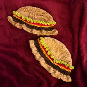 May include: Two handmade crochet hats in a taco design. The hats are tan with brown, red, and yellow stripes, and a green wavy trim. The hats are on a burgundy background.