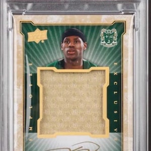 May include: A Lebron James trading card from 2015, encased in a protective holder. The card features a photo of Lebron James, a piece of memorabilia, and his autograph. The card is graded GEM MT 10 by PSA.