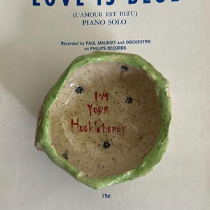 Op de afbeelding: Een handgemaakte keramische kom met een gespikkeld beige interieur en een groene rand. De kom is beschreven met de woorden "I'M YOUR HUCKLEBERRY" in het rood. De kom staat op een vintage bladmuziek achtergrond.