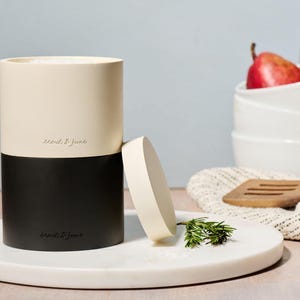 May include: A two-tone kitchen utensil holder with a cream top and black base, with the text "leeds & june". The lid is off to the side. A marble tray holds the holder, rosemary, and a wooden spatula.
