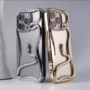 Pode incluir: Duas capas de telefone, uma prateada e outra dourada, cada uma com um design de cobra. A cobra envolve as bordas do telefone, adicionando um elemento decorativo. As capas são exibidas em um fundo neutro.