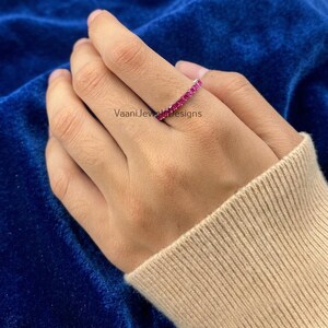 May include: A silver ring featuring a continuous row of square-cut, bright pink gemstones. The ring is displayed on a hand, with the brand name "VaaniJewelDesigns" visible. The background is a dark blue fabric, and the sleeve is beige.