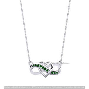 May include: A silver-toned necklace featuring an infinity symbol intertwined with a heart shape. The pendant is accented with small, round, green stones set along the curves of the design. The piece includes a delicate chain and the text VaaniJewelsDesigns.