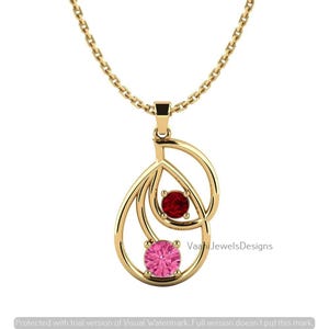 May include: A gold pendant necklace featuring an open teardrop frame with two round gemstones. The upper stone is a deep red, while the lower stone is a bright pink. The pendant hangs from a delicate gold chain, and the text VaahiJewelsDesigns is visible.