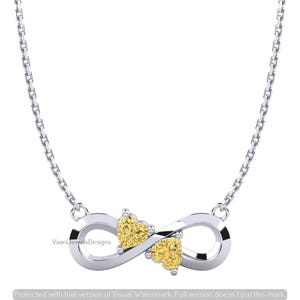 May include: A silver-toned necklace featuring an infinity symbol pendant adorned with two yellow heart-shaped gemstones. The pendant is attached to a delicate silver chain. The text VaaniJewelsDesigns is visible on the left side of the infinity symbol.