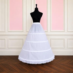 May include: A white, tiered petticoat with a ruffled hem is displayed on a black mannequin. The petticoat has three horizontal bands and is set against a pink and white backdrop. The skirt is designed to add volume to a dress.
