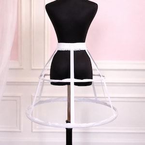 Puede incluir: Un miriñaque blanco sobre un maniquí negro. El miriñaque tiene un diseño circular estructurado con aros y una cintura blanca. El maniquí se encuentra sobre un fondo rosa y blanco.