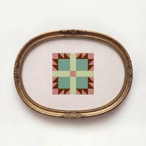 May include: A cross-stitch embroidery featuring a geometric quilt block pattern in shades of dusty pink, teal, light green, and dark brown, centered on white Aida fabric and enclosed within an ornate, oval-shaped metallic gold frame.