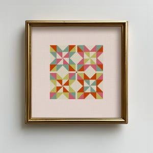 May include: A framed cross-stitch artwork featuring a geometric pattern of colorful triangles. The design includes orange, pink, green, and blue shapes arranged in a repeating star-like pattern. The artwork is set in a gold frame.