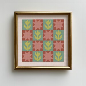 Puede incluir: Una obra de arte en punto de cruz enmarcada con un patrón repetido de flores amarillas estilizadas y formas de estrella en tonos de rojo, verde y marrón. La obra de arte está sobre un fondo rosa claro y enmarcada en un marco dorado.