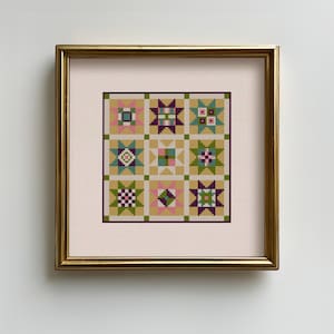 May include: A framed cross-stitch sampler featuring a patchwork quilt design. The quilt blocks are in shades of green, pink, purple, and gold, set against a cream background. The frame is gold.