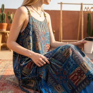 May include: A blue patterned maxi dress with a paisley and floral design. The dress has spaghetti straps with green bead accents and a drawstring waist. The person is holding a white paper cup with a black lid.