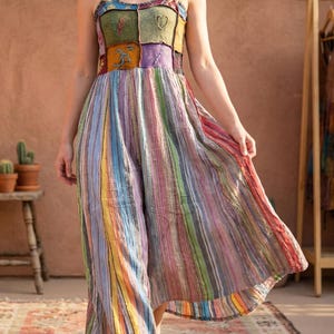 Op de afbeelding: Een kleurrijke, gestreepte jumpsuit met een patchwork lijfje. Het lijfje heeft vierkanten in verschillende kleuren en geborduurde details. De jumpsuit heeft wijde pijpen en een vloeiend ontwerp, met verticale strepen in tinten rood, geel, blauw en groen.