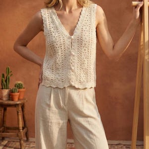 May include: Cream-colored crochet top with a V-neck and button closure. The sleeveless top has a scalloped edge and is paired with light beige linen pants. The outfit is styled in a neutral color palette.