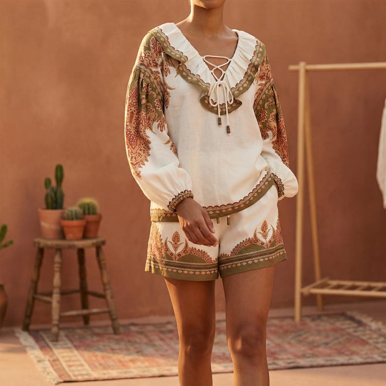 May include: White blouse and shorts set with a paisley pattern in shades of green and rust. The blouse has long sleeves, a V-neck with a lace-up detail, and a ruffled collar. The shorts have a matching patterned border.