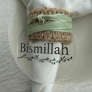 Könnte beinhalten: Eine transparente, runde Tischkarte mit dem Wort "Bismillah" in schwarzer Schrift, verziert mit einem zarten floralen Design. Die Karte liegt auf einer weißen Serviette, gehalten von einem Serviettenring aus Jute und grünem Band.