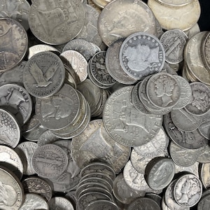 May include: A collection of silver coins, including quarters, dimes, and half dollars, are scattered across a dark surface. The coins display various designs and dates, showcasing historical American currency. The image highlights the details of each coin's engravings.