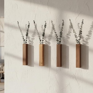 Peut inclure: Quatre vases muraux en bois, chacun avec une teinte unique, sont montés sur un mur blanc texturé. Chaque vase contient une brindille de verdure avec de petites feuilles sombres. Les vases sont rectangulaires avec un design géométrique.