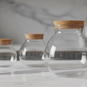 May include: Three clear glass jars with cork lids of varying sizes are arranged on a white surface. The jars have a rounded shape and are empty. The background includes a blurred kitchen setting with a marble countertop and wooden cutting board.