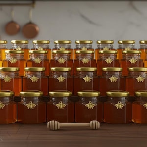 Puede incluir: Una pirámide de frascos hexagonales de vidrio llenos de miel dorada, cada uno con una tapa dorada y un emblema de abeja. Un cucharón de miel de madera descansa frente a los frascos. Los frascos están dispuestos sobre una superficie de madera.