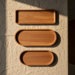 Puede incluir: Tres bandejas de madera con un acabado liso de color marrón claro. La bandeja superior es rectangular con esquinas redondeadas, mientras que las bandejas central e inferior son ovaladas. Cada bandeja tiene un centro poco profundo y bordes elevados.