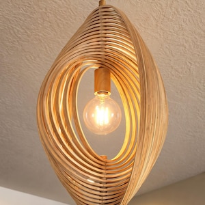 May include: A pendant light fixture featuring a series of curved, light-colored wooden slats arranged in an oval, cage-like structure. A single round light bulb is suspended in the center, attached to a brass-colored metal rod and ceiling mount.