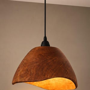 May include: A pendant light fitting featuring a curved, bowl-shaped shade made of dark brown wood with a visible grain pattern. The shade is suspended by a thin black cord from a matching circular wooden ceiling canopy, with a warm light glow.