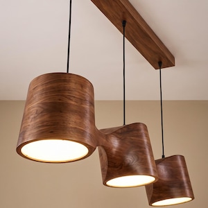 May include: A ceiling light fixture featuring three cylindrical wooden shades with a natural grain pattern, suspended by black cords from a rectangular wooden mounting plate. Each shade emits a warm, downward-facing light from its circular base.