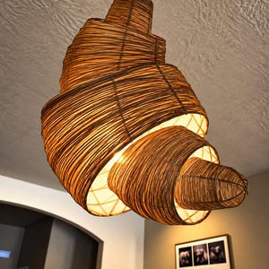 Può includere: Una lampada a sospensione realizzata con strisce di vimini marrone avvolte, che formano una spirale a forma di conchiglia. La struttura presenta un design a trama aperta che permette alla luce calda di uscire dall'interno, sospesa da un cavo nero.