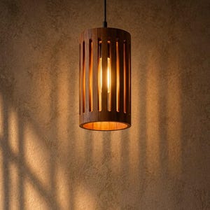 Könnte beinhalten: Eine zylindrische Pendelleuchte aus dunklem Holz mit vertikalen Lamellen. Die Lampe ist an einem dünnen schwarzen Kabel an einer passenden runden Deckenhalterung aus Holz befestigt. Im Inneren des Holzrahmens ist eine warm leuchtende Glühbirne.