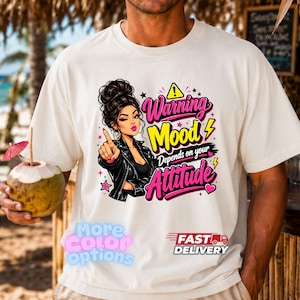 Pode incluir: Uma t-shirt de cor creme com o gráfico de uma pessoa com um penteado de coque alto e um casaco de cabedal preto. O texto diz Warning Mood Depends on your Attitude, acompanhado por raios amarelos, estrelas cor-de-rosa e corações.