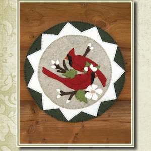 May include: A felt applique pattern for a 12 1/2 inch diameter round wall hanging featuring two red cardinals perched on a branch with white blossoms. The pattern is titled "Cardinals & Blooms" and is from Cotton Tales Patterns.