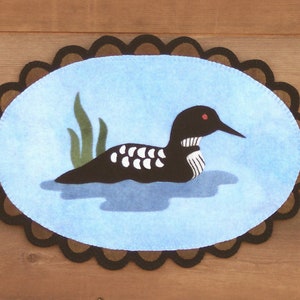 May include: A felt wall hanging with a brown scalloped border. The centre of the hanging is a light blue background with a black and white loon swimming in the water. The loon has a red eye and is surrounded by green reeds.
