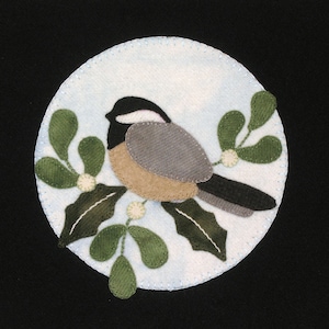 Op de afbeelding: Een vilt applique van een zwartkopmees die op een tak mistel met groene bladeren en witte bessen zit. De vogel is bruin, grijs en zwart. De achtergrond is een lichtblauwe cirkel met een witte rand.